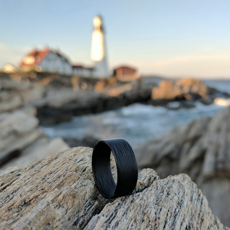 Ultralight Men's Ring with Wave Carbon Fiber On The Rocks Of A Beach In Front Of A Lighthouse